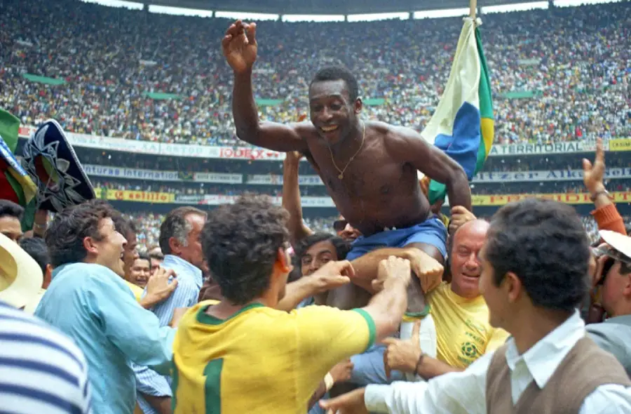 A soccer player, shirtless and smiling, is lifted on the shoulders of teammates and fans in a crowded stadium celebrating, with a Brazilian flag visible in the background.