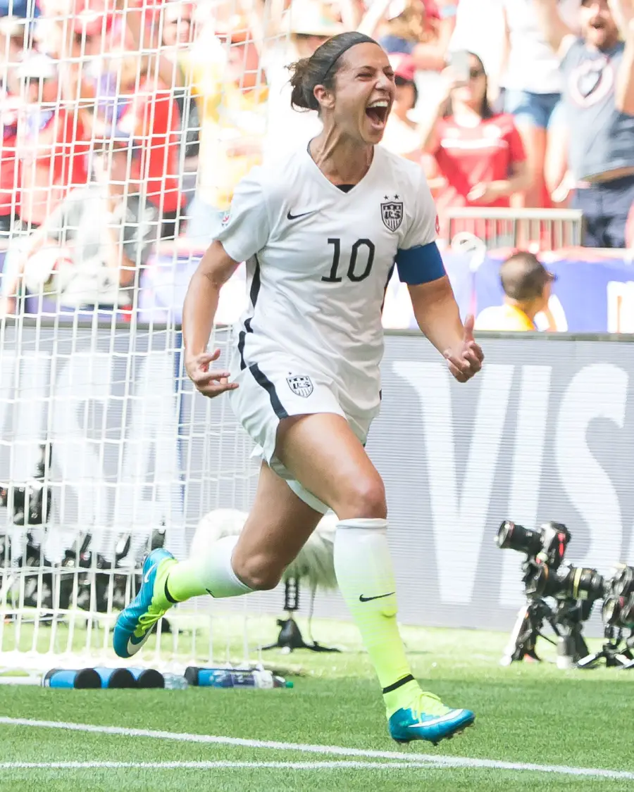 A soccer player in a white USA jersey with the number 10 celebrates passionately on the field, mouth open and fists clenched, with fans and photographers in the background.