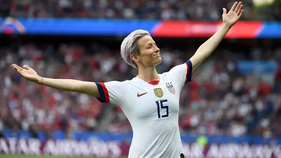 A soccer player wearing a white USA jersey with the number 15 stands on the field with arms outstretched, smiling as a crowd watches from the stands in the background.