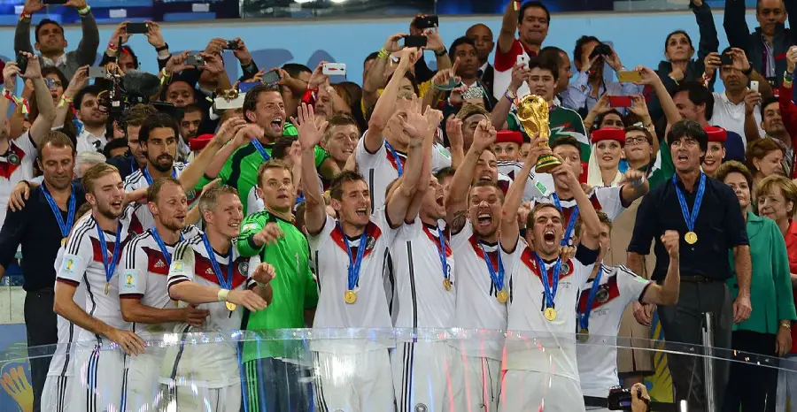 A soccer team in white jerseys celebrates on a stage, holding up a golden trophy as fans cheer and take photos. Many players wear gold medals, and confetti is falling around them.