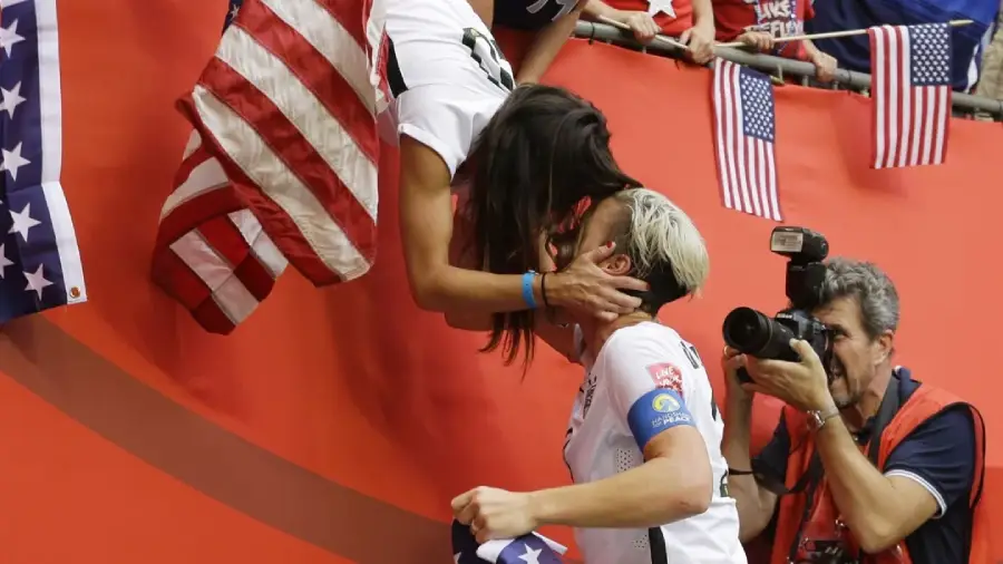 Two soccer players celebrate by sharing a kiss over a barrier, surrounded by American flags, while a photographer captures the moment. The scene is filled with excitement and national pride.