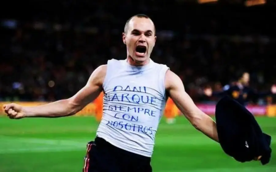 A soccer player celebrates passionately on the field, wearing a sleeveless shirt with a handwritten message in Spanish. He is shouting with one arm outstretched and holding a dark shirt or jersey in the other hand.