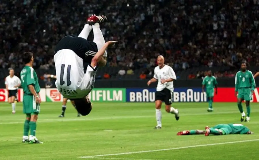 A soccer player in a white jersey does a mid-air backflip to celebrate a goal, while other players and a goalkeeper in green react on the field during a crowded stadium match.