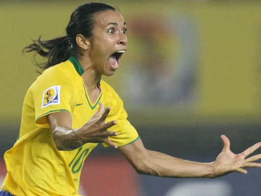 A Brazilian soccer player in a yellow jersey with number 10 celebrates passionately on the field, mouth wide open and arms outstretched, showing excitement and energy.