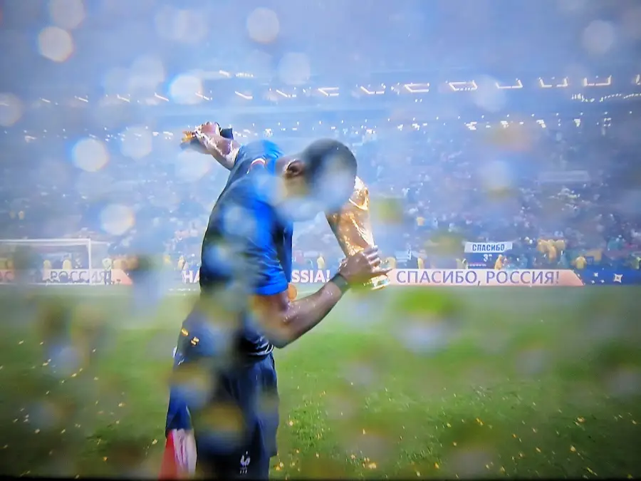 A soccer player in a blue jersey bows his head while holding up a gold trophy in a rainy stadium, with water droplets on the camera lens and a cheering crowd in the background.
