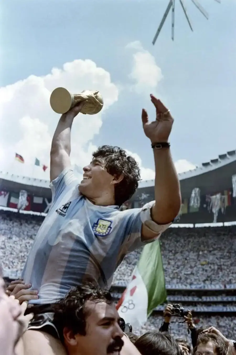 A soccer player in a light blue and white jersey is lifted on shoulders, holding a gold trophy and waving amid a cheering crowd in a large stadium.