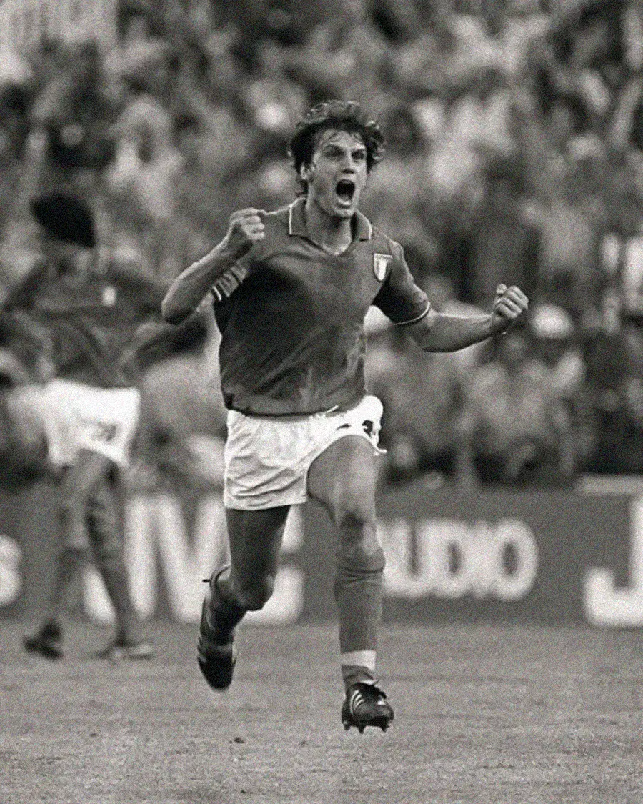 A soccer player in an Italy jersey celebrates with clenched fists, running on the field. The black-and-white photo shows fans and another player in the background, capturing a moment of excitement during a match.