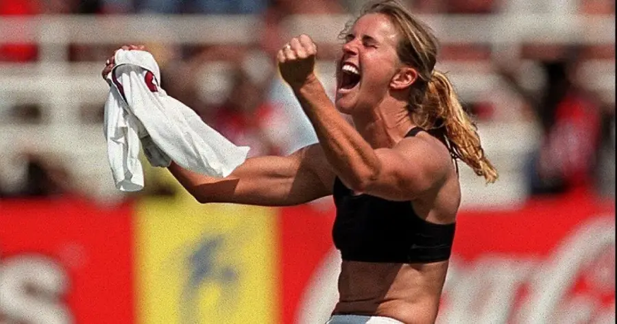A female soccer player celebrates passionately on the field, clenching her fist and holding her shirt in one hand, wearing a black sports bra, with a stadium crowd blurred in the background.