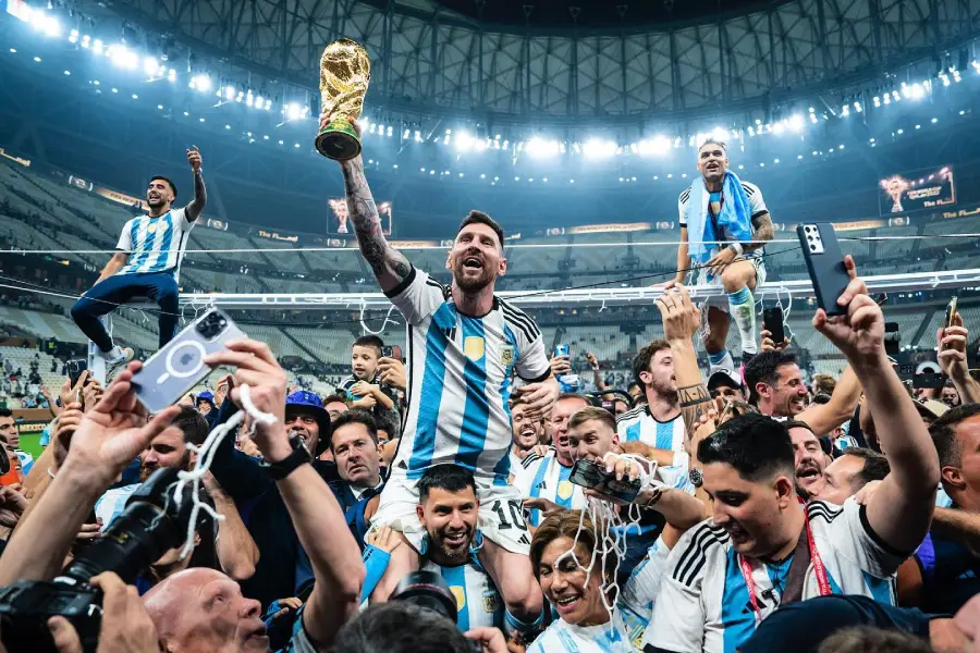 A soccer player in an Argentina jersey holds the World Cup trophy aloft while surrounded by jubilant teammates and fans in a stadium, celebrating their victory.