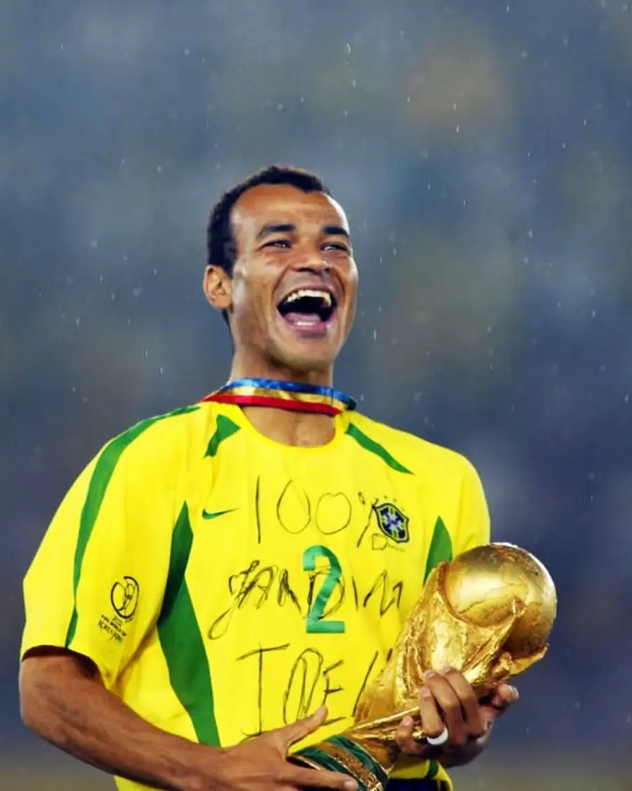 A smiling soccer player in a yellow Brazil jersey holds the FIFA World Cup trophy, celebrating on a rainy field.