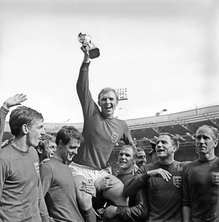 A football player in an England jersey is lifted by teammates as he holds a trophy aloft in a stadium. Other teammates cheer and smile around him, celebrating the victory. The image is in black and white.