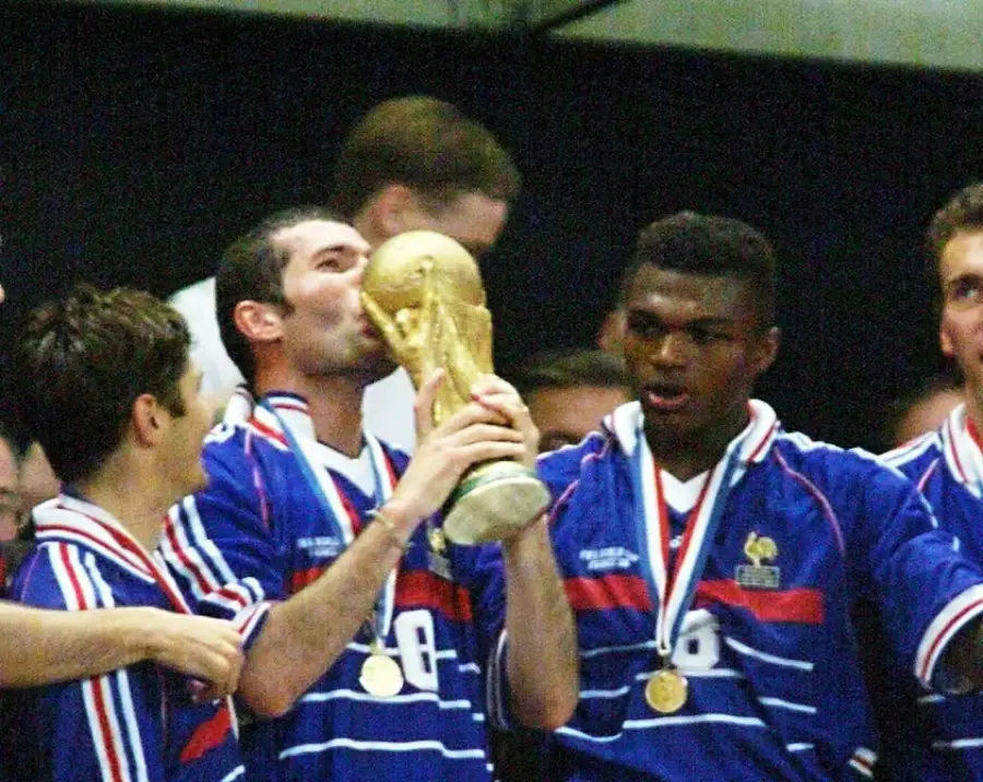 French soccer players celebrate winning the World Cup. One player kisses the trophy while others smile and wear gold medals, all dressed in blue France jerseys.