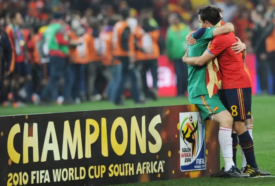 Two soccer players hug near a sign that reads "CHAMPIONS 2010 FIFA WORLD CUP SOUTH AFRICA" on a field, celebrating after a match with people and photographers in the background.