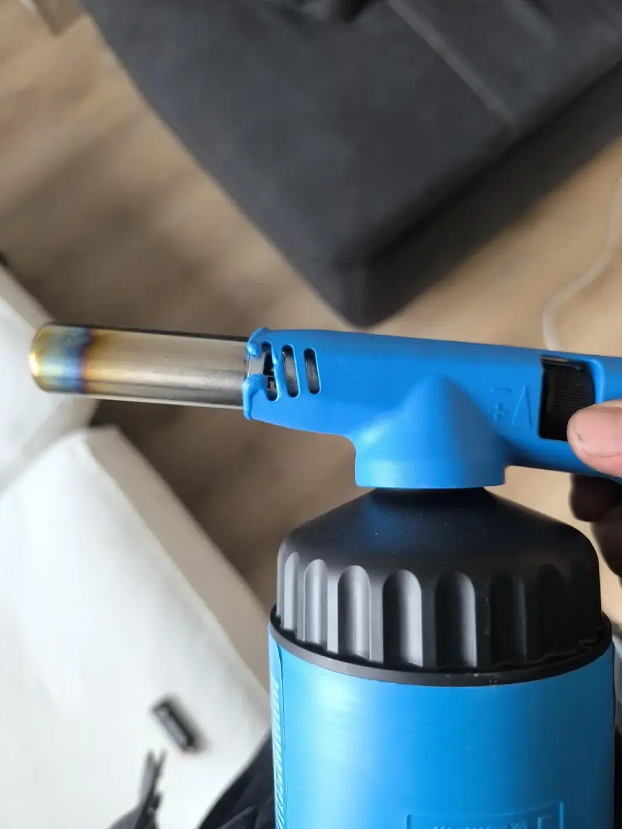 A close-up of a hand holding a blue blowtorch with a black base, showing the metal nozzle with burn marks. A couch and wooden floor are blurred in the background.