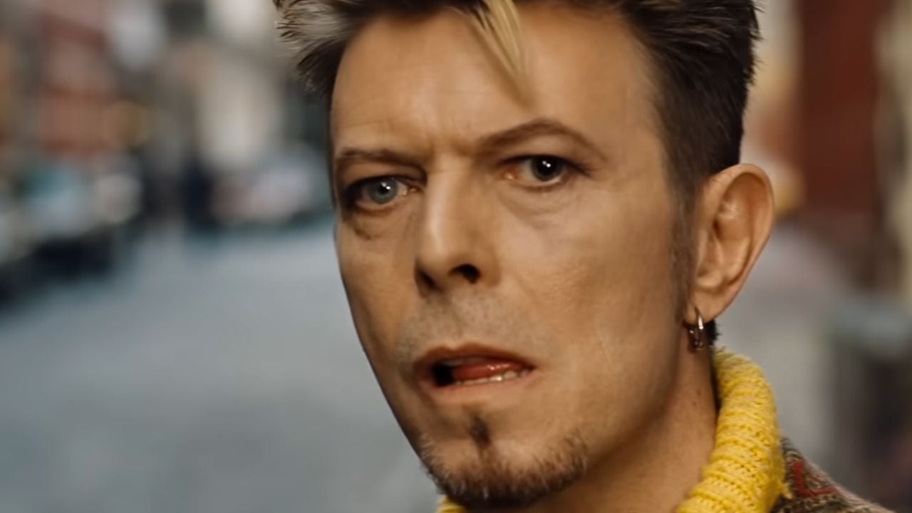 A person with light skin, styled blond and brown hair, and a short goatee looks ahead with a neutral expression. They wear a yellow sweater and an earring, with a blurred city street in the background.
