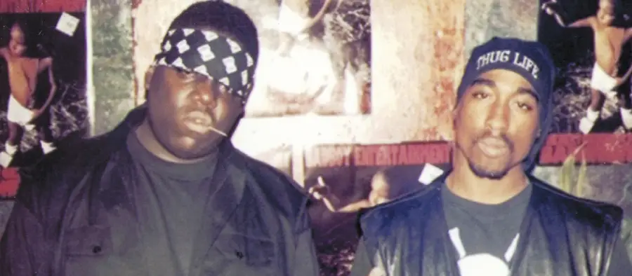 Two men stand side by side. The man on the left wears a bandana over his head and dark clothes; the man on the right wears a "Thug Life" beanie and a vest. Posters with a baby are visible in the background.