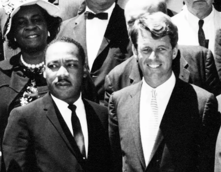 Two men in suits stand side by side, smiling, among a group of people dressed formally; one man wears a tie and the other a dark suit and tie.