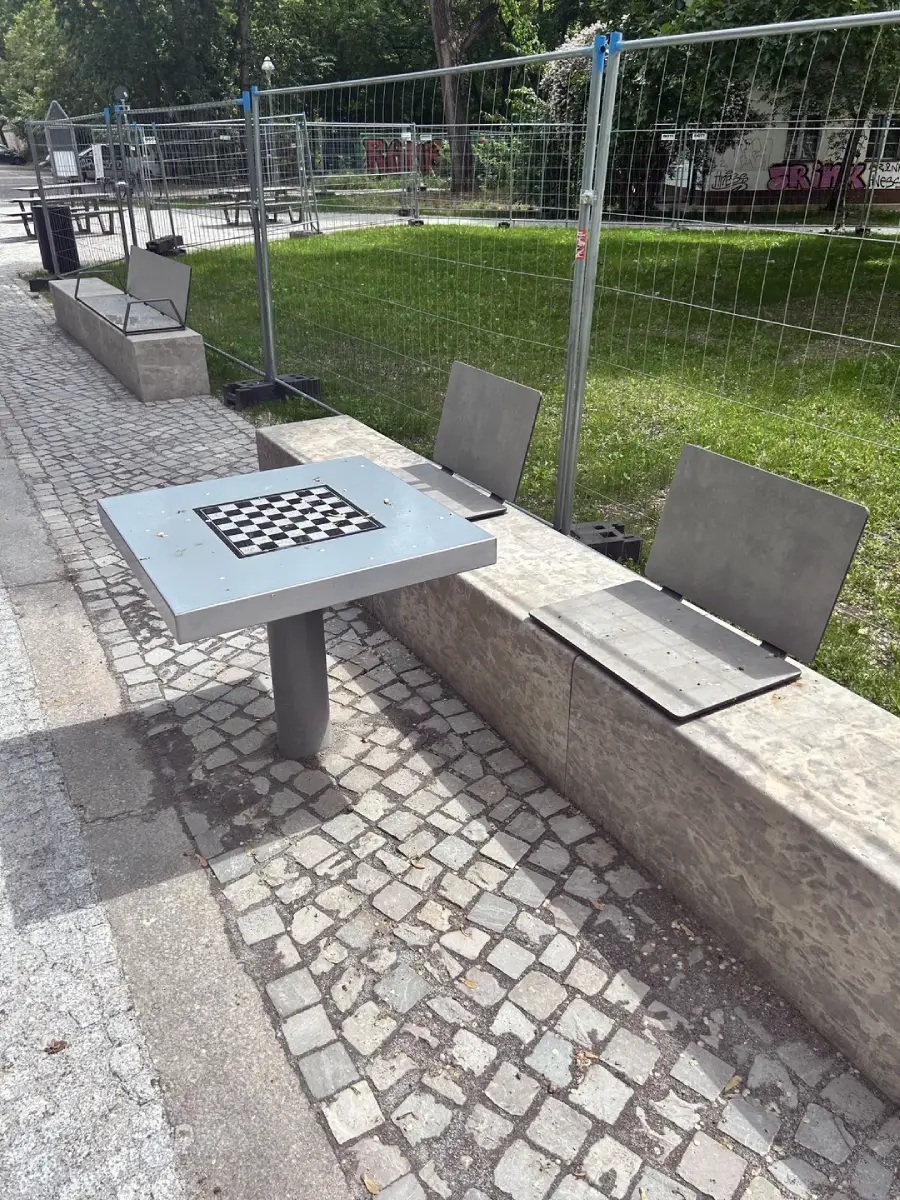 Two metal benches with missing seat sections face a chess table on a stone sidewalk. There is a fenced grassy area and trees in the background, suggesting a park or public space.