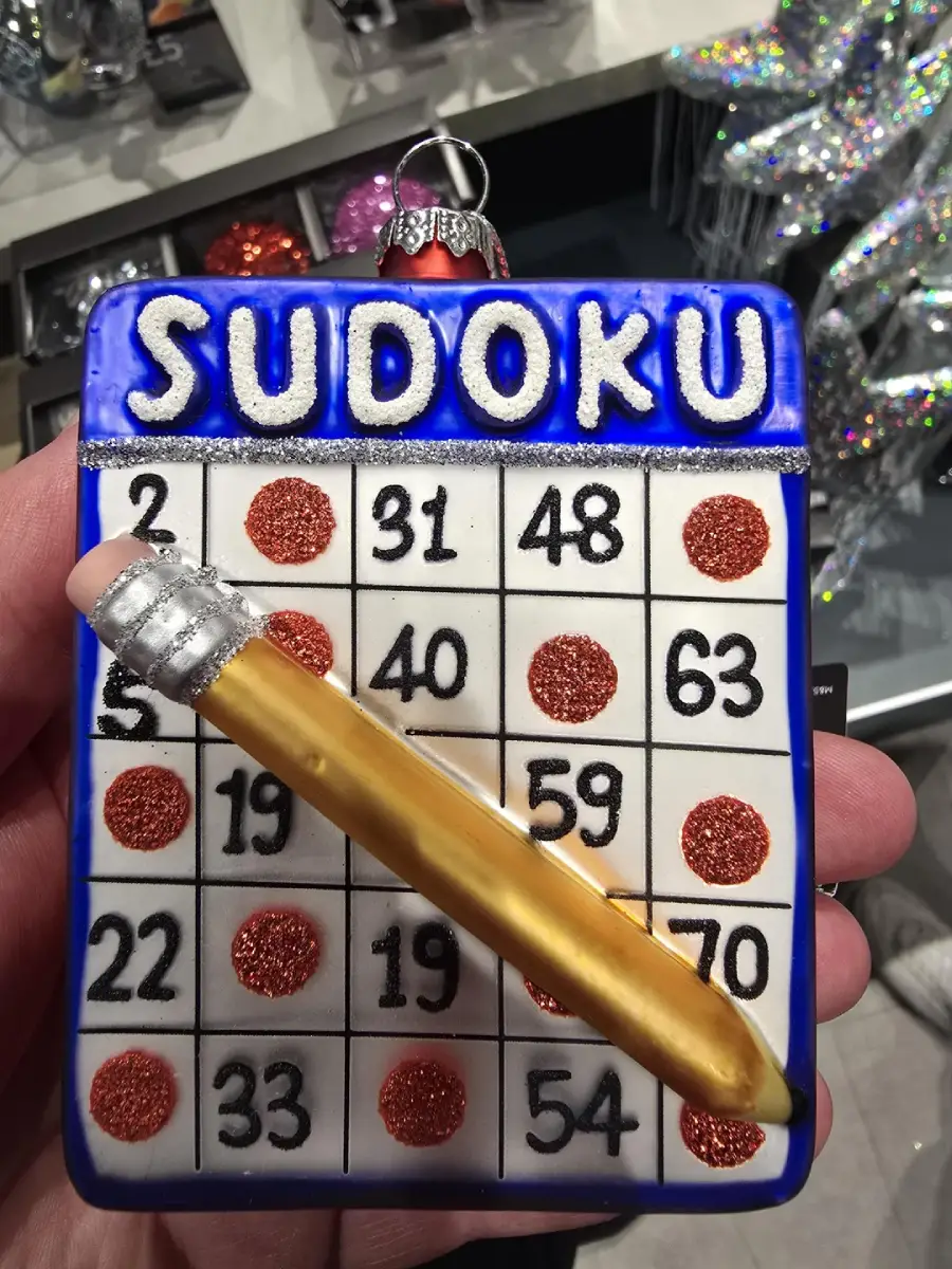 A hand holds a decorative ornament resembling a sudoku puzzle board with glittery red circles and a large yellow pencil attached diagonally across it. The word "SUDOKU" is written in bold white letters at the top.