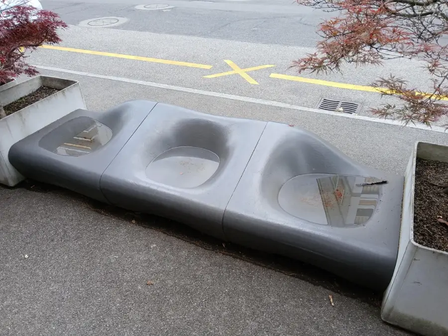 A modern, curved gray bench with three indented seats collects rainwater in each seat, making them unusable. The bench is placed on a sidewalk between two large square planters. A road is visible in the background.