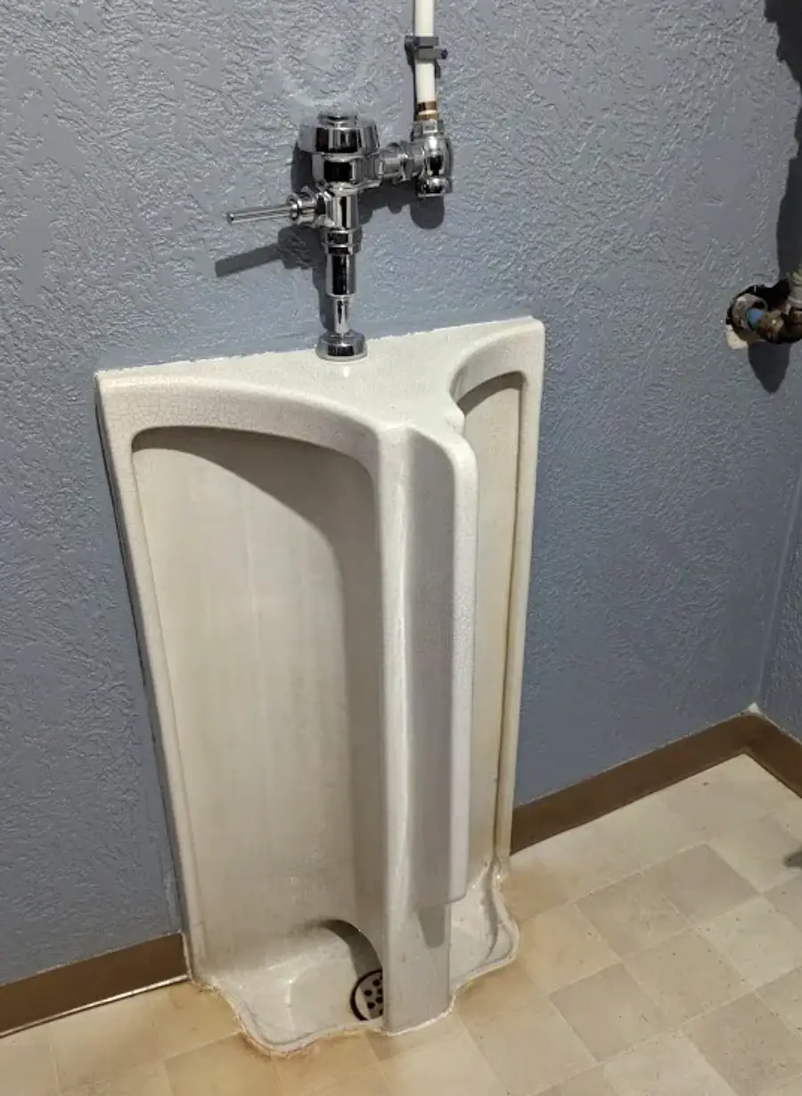A white urinal with an unusual divider down the middle is installed against a textured gray wall with a metal flush valve above it and tan baseboards along the floor.