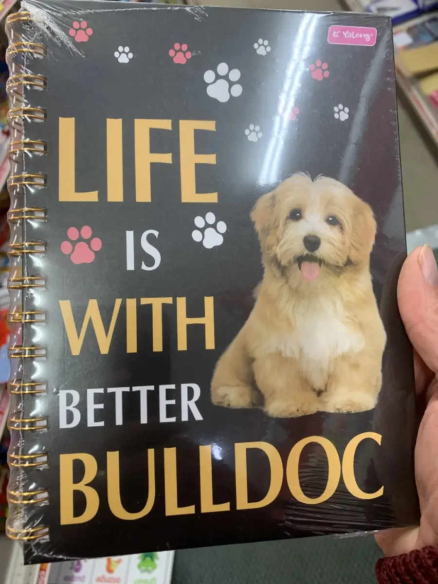 A hand holds a spiral-bound notebook with a cute fluffy dog on the cover. The text reads, "Life is better with bulldoc," with paw prints decorating the black background. The dog does not resemble a bulldog.