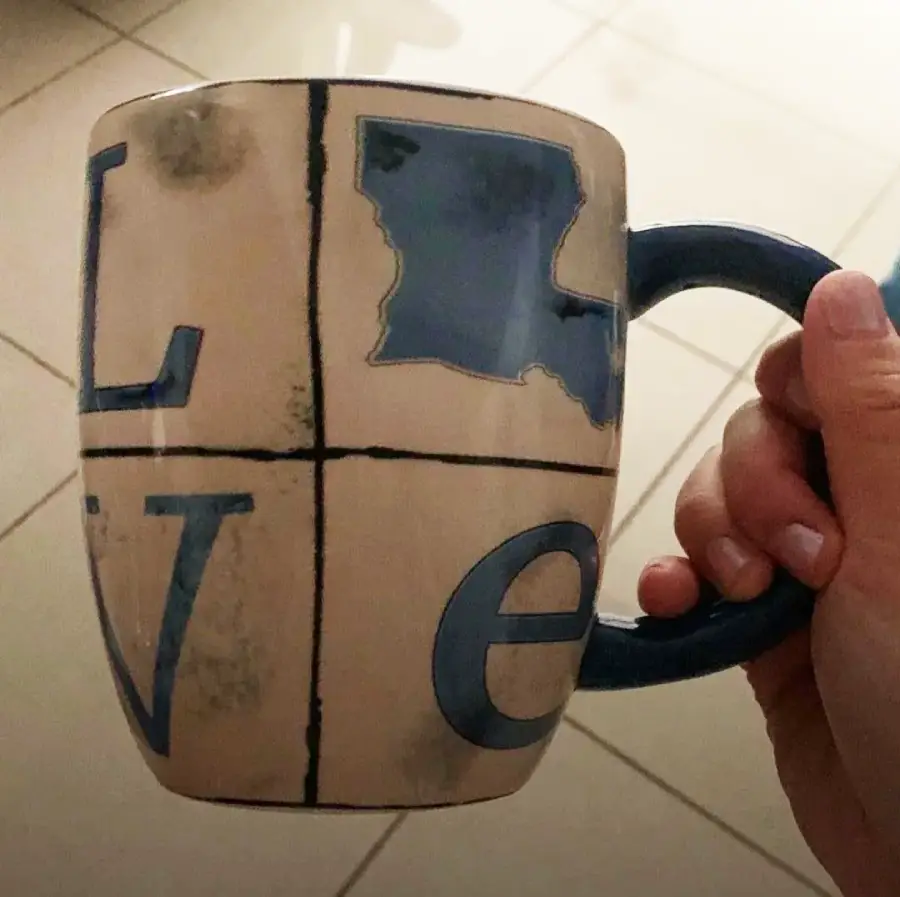 A hand holds a mug with the word "love," where the "o" is replaced by the outline of the state of Louisiana. The design is in shades of blue and beige, and tile flooring is visible in the background.