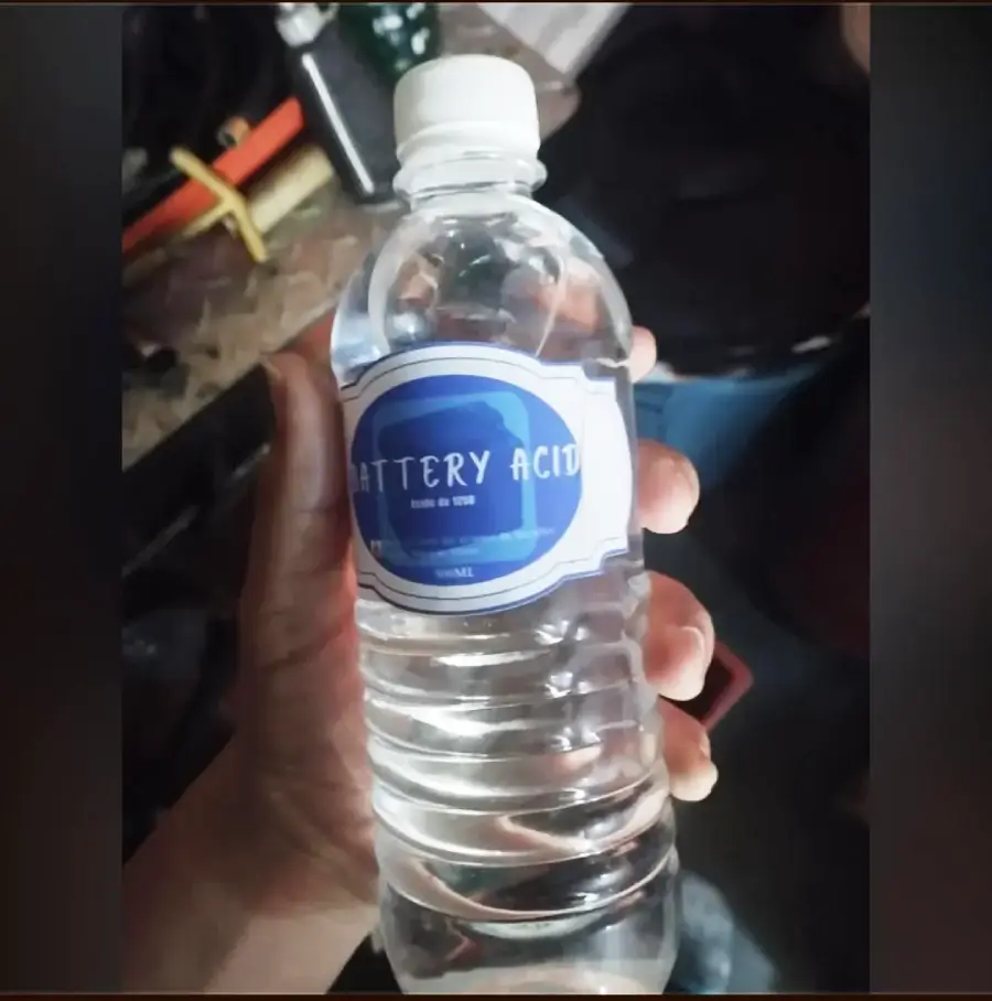 A hand holds a clear plastic water bottle labeled "Battery Acid," though the bottle appears to be filled with water. The background shows a cluttered surface with various objects.