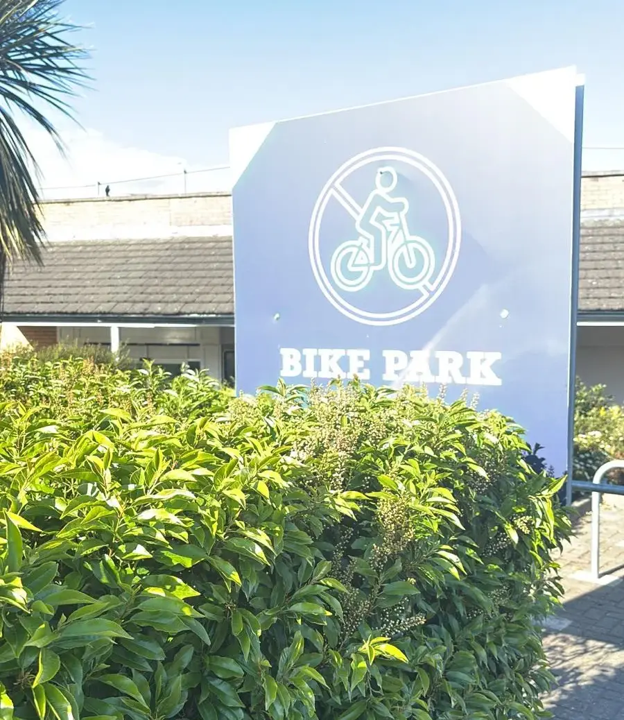 A sign reading "Bike Park" with a bicycle symbol stands behind a green hedge, with a building and parts of a palm tree visible in the background under a clear sky.