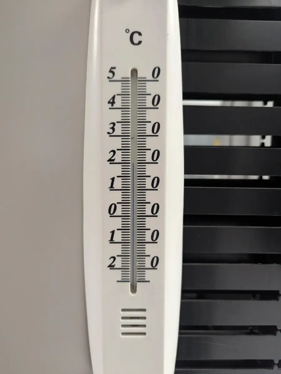 A close-up of a white thermometer showing temperatures in degrees Celsius, ranging from -30°C to 50°C. The current temperature appears to be about 27°C. The background has a black vented surface.