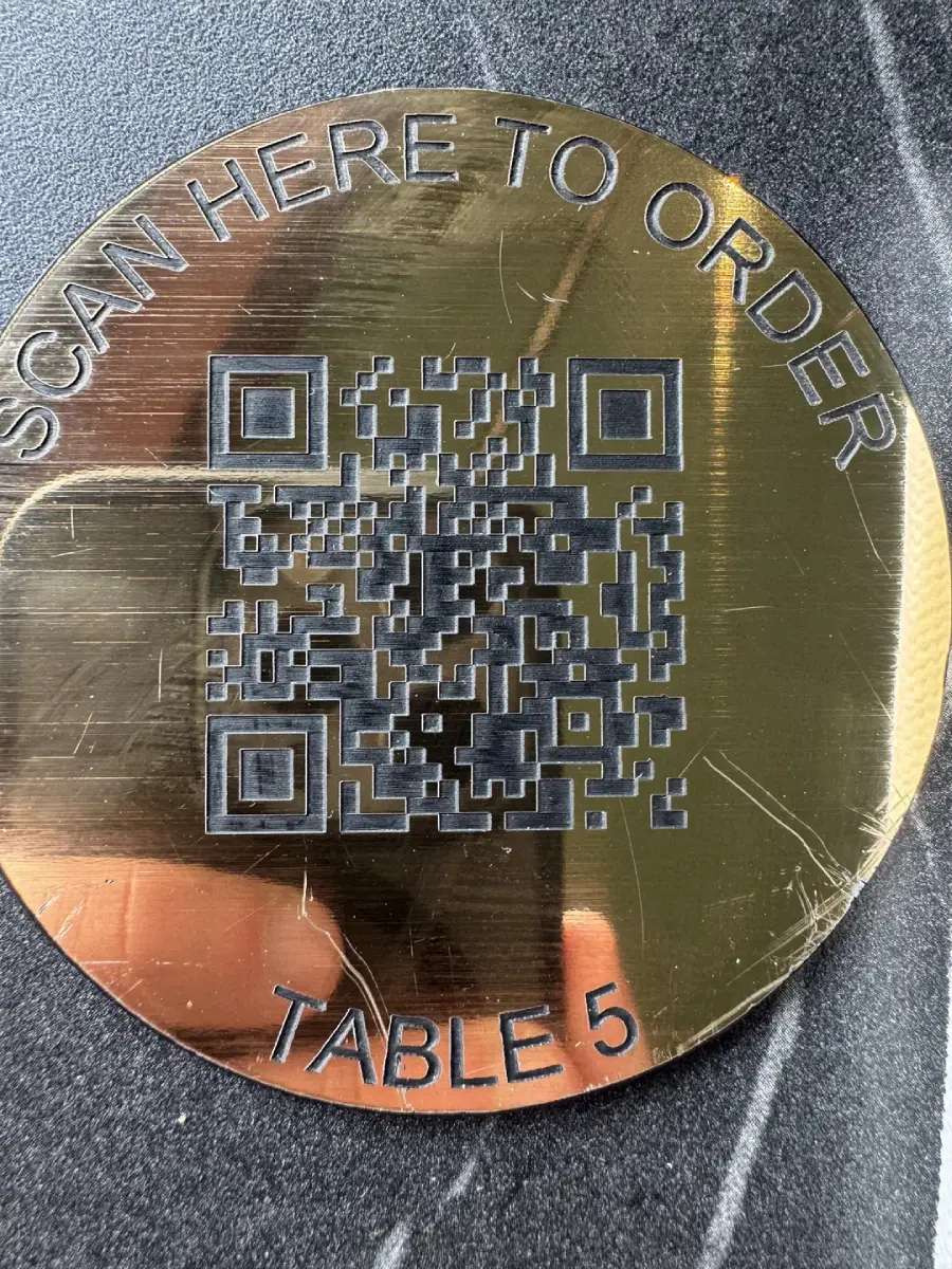 A round metallic plaque on a dark surface features a QR code in the center with the words "SCAN HERE TO ORDER" above and "TABLE 5" below.