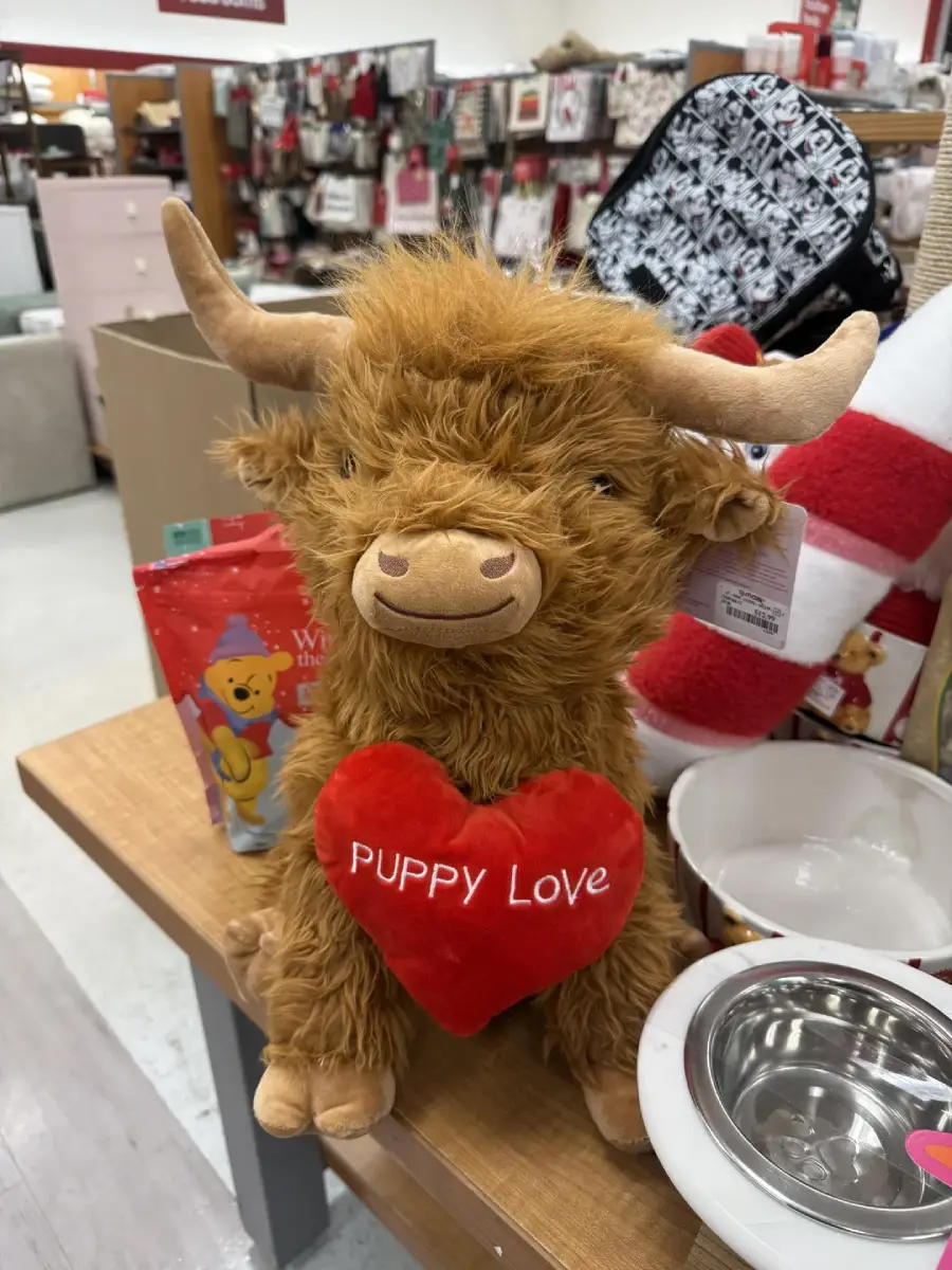 A plush highland cow with tan fur and horns holds a red heart-shaped pillow that says "PUPPY LOVE." The toy is on a store display table among various pet and holiday items.