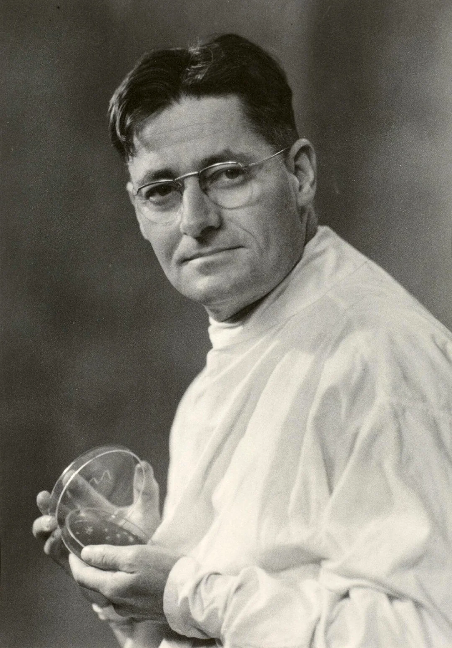 A man wearing glasses and a white lab coat holds a petri dish, looking slightly to the side. The image is in black and white, with a soft, blurred background.