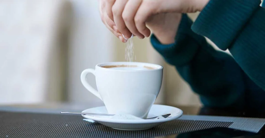 A person sprinkles sugar into a white cup of coffee on a saucer with a spoon, sitting at a table.