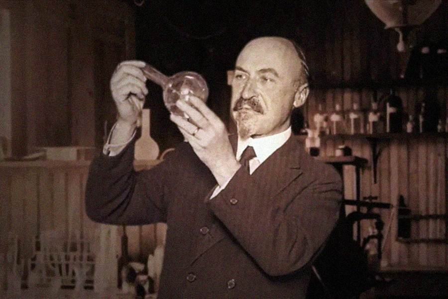 A man in a suit holds up a round-bottom flask and examines it in a vintage laboratory, surrounded by shelves with glassware and scientific equipment.