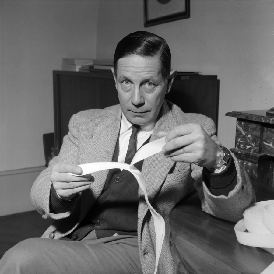A man in a suit sits at a desk, holding a white strip of paper and looking intently at the camera. There are books and a wooden cabinet in the background.