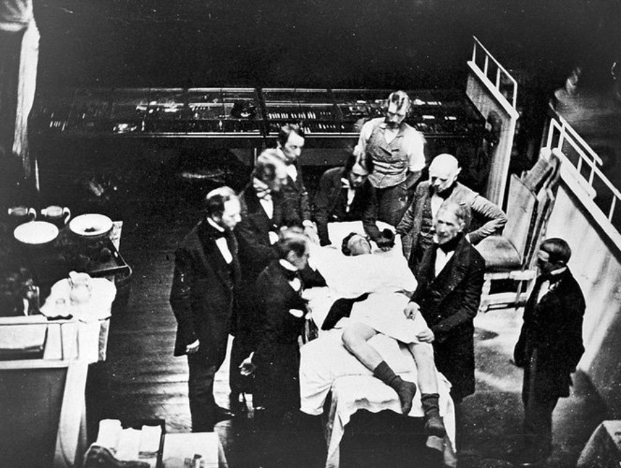 A black and white photo showing several men in suits gathered around a patient lying on a hospital bed in an operating room, observing a medical procedure or demonstration.