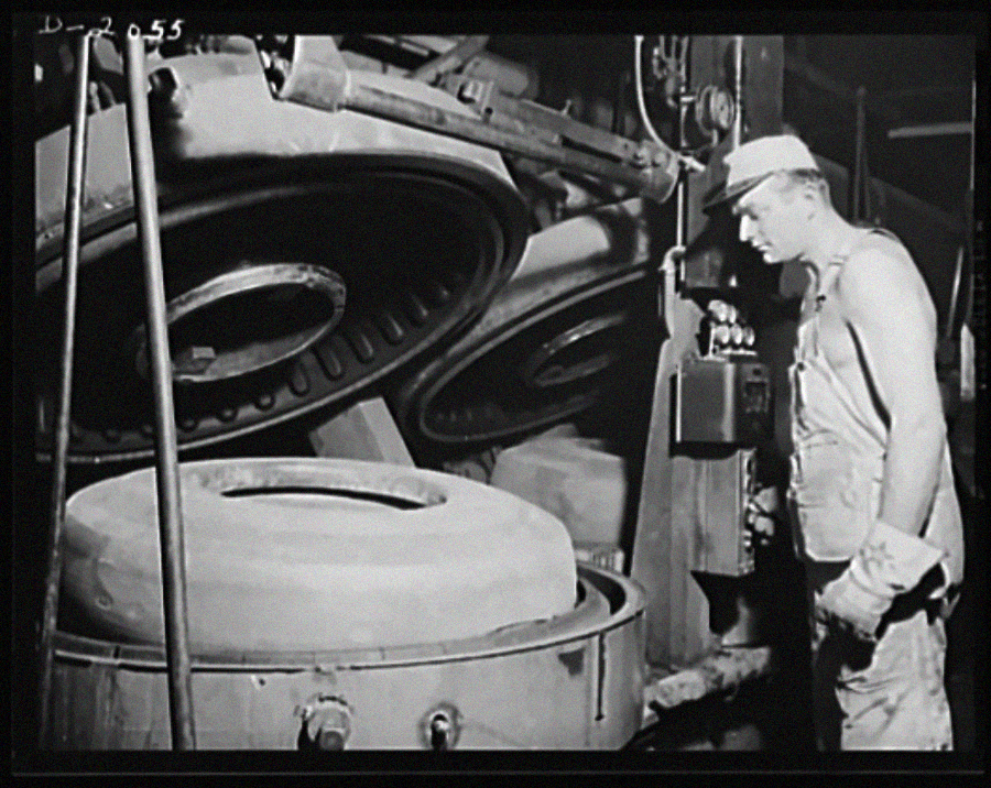 A man in work clothes and gloves stands near large industrial machinery, closely observing a round mold or part being processed inside a manufacturing facility.