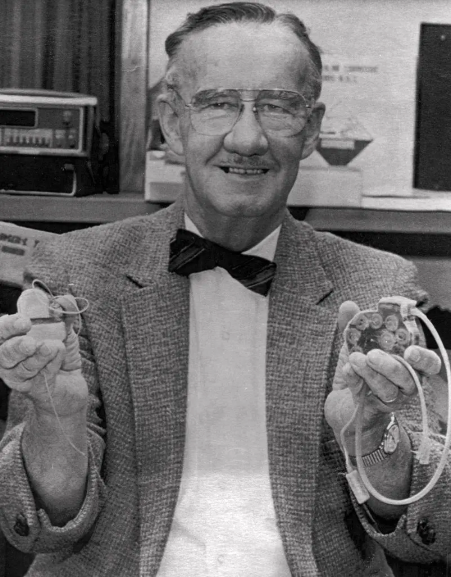 An older man wearing glasses, a bow tie, and a suit jacket smiles while holding two early hearing aid devices, one in each hand, in what appears to be an office or laboratory setting.