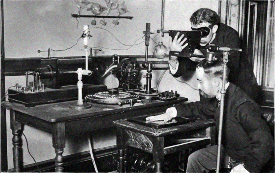 Two men in early 20th-century attire operate a collection of vintage scientific equipment; one sits at a table with various devices while the other looks through a stereoscope-type viewer.