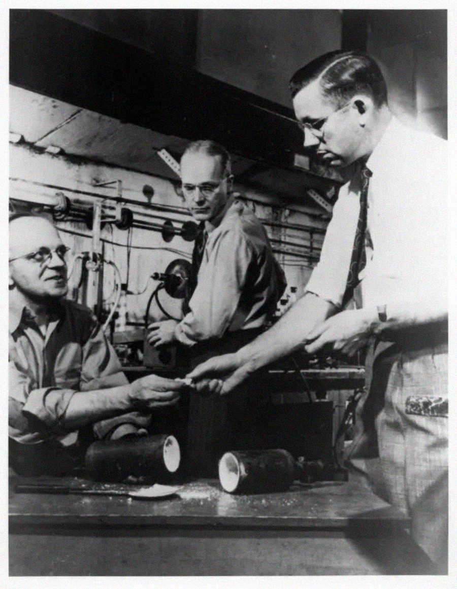 Three men work together in a laboratory, with one man handing an object to another while the third observes. Laboratory equipment and cylindrical objects are on the table in front of them.