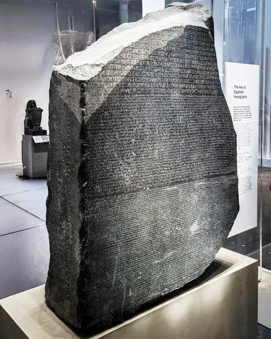 The Rosetta Stone, a large, dark stone slab with inscriptions in three scripts, displayed upright in a glass case at a museum. Informational plaques and other artifacts are visible in the background.
