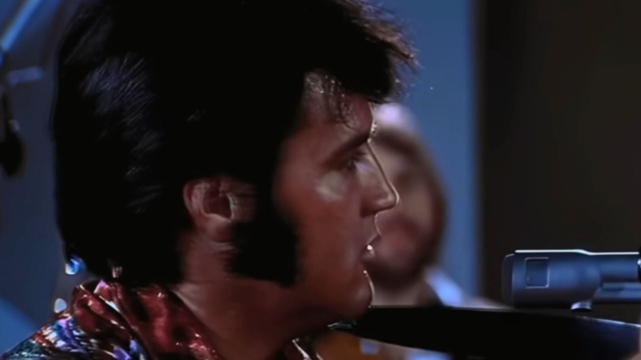 A man with dark hair and sideburns sings into a microphone. He wears a colorful shirt and is shown in profile, with a blurred figure in the background.