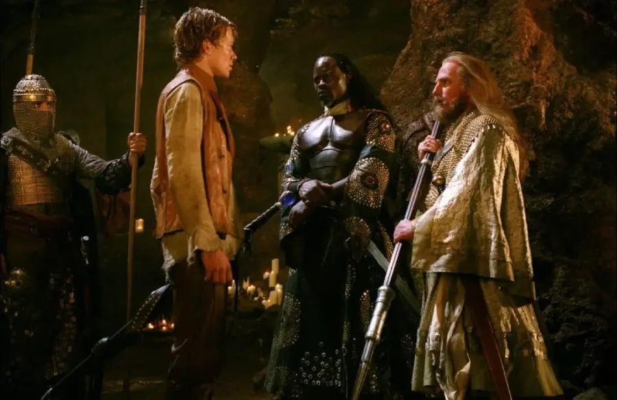 Four people in medieval-style costumes stand in a dimly lit cave, surrounded by candles. One wears armor, two hold staffs, and they appear to be in serious conversation.