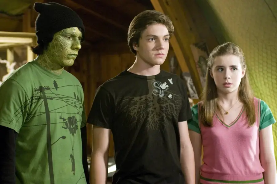 Three teenagers stand indoors. The one on the left has green, textured skin and wears a beanie and green shirt. The middle teen wears a black shirt with a white design, and the girl on the right has long hair and a pink top.