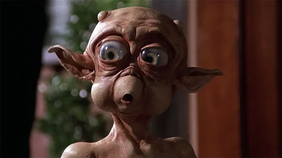 A close-up of a wide-eyed alien character with large ears, wrinkled skin, and an expressive, surprised face. The background is blurred with hints of greenery and wood.