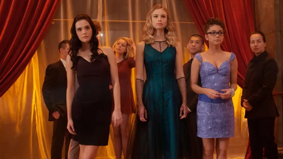 A group of young adults in formal attire stand together in front of red curtains. Three women in elegant dresses are at the center, flanked by people in dark suits and evening wear, with warm lighting in the background.