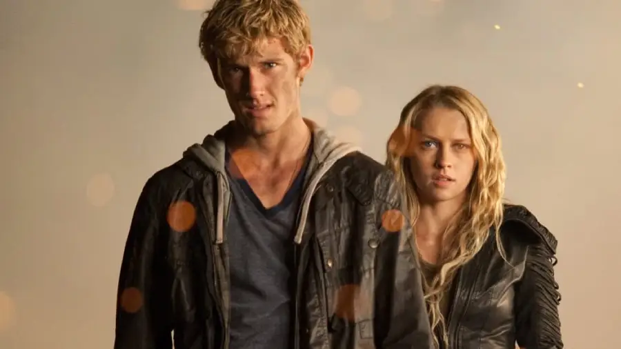 A young man and woman with serious expressions stand side by side, both wearing dark jackets against a neutral, softly lit background.