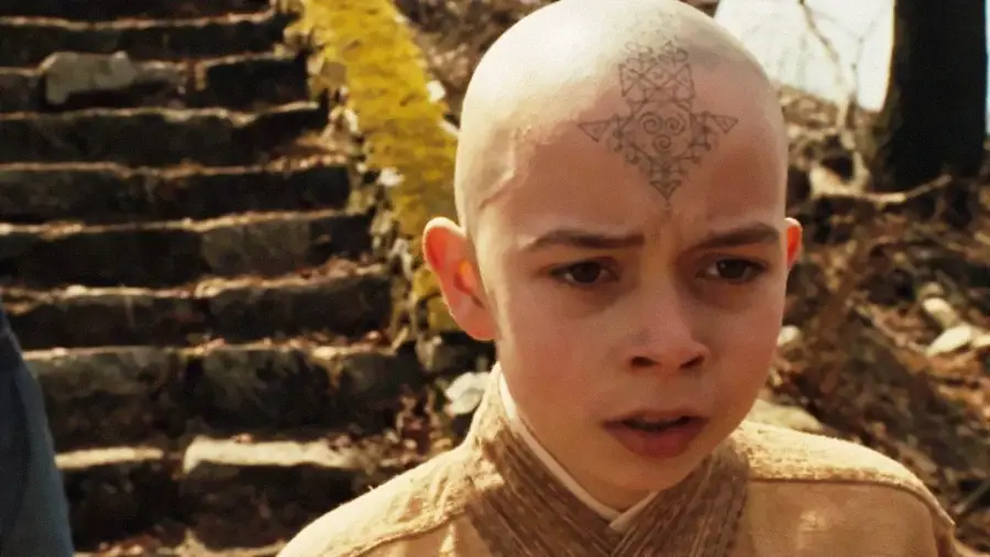 A young boy with a shaved head and a blue arrow tattoo on his forehead stands outdoors near stone steps, wearing a tan, robe-like outfit. He appears concerned or focused.