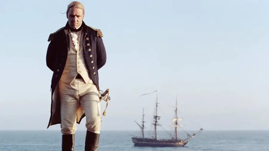 A man in an 18th-century naval uniform stands on a ship’s deck with his hands behind his back. In the background, a large sailing ship floats on the open sea under a clear sky.